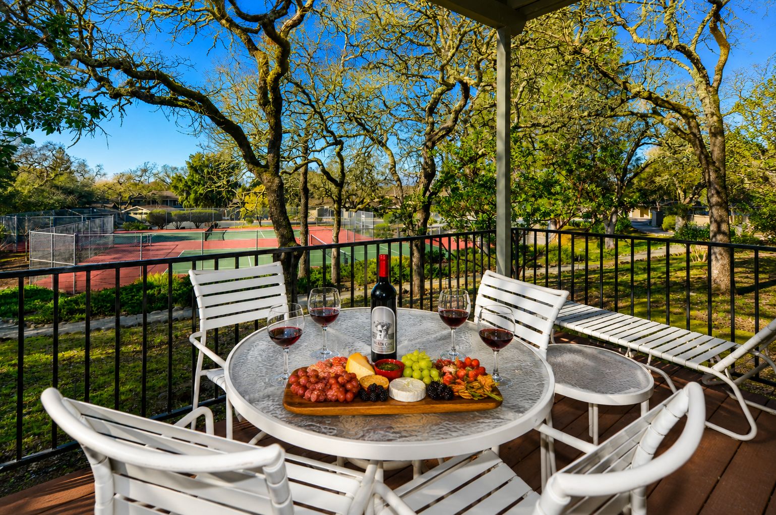 An outdoor dining area with a table set for two, surrounded by white chairs, a tennis court in the background, and trees.