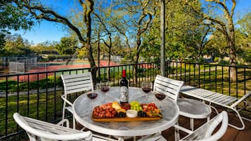 An outdoor dining area with a table set for two, surrounded by white chairs, a tennis court in the background, and trees.
