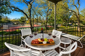 An outdoor dining area with a table set for two, surrounded by white chairs, a tennis court in the background, and trees.