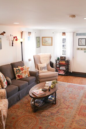 A cozy living room with a sofa, armchair, and coffee table, featuring a rug, framed artwork, and a bookshelf.