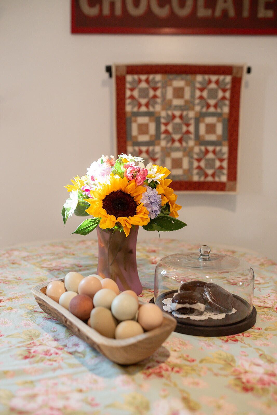 A table with a floral tablecloth, a vase with flowers, a bowl of eggs, and a glass dome over a chocolate dessert.