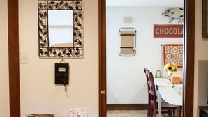 A hallway with a wooden floor, a wall-mounted mirror, a small table with a bowl and a candle, and a door leading to a dining area with a table and chairs.
