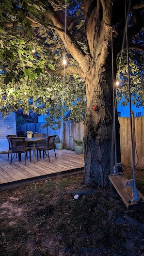Eine Holzterrasse mit Tisch und Stühlen, umgeben von einem Baum und einer Schaukel.