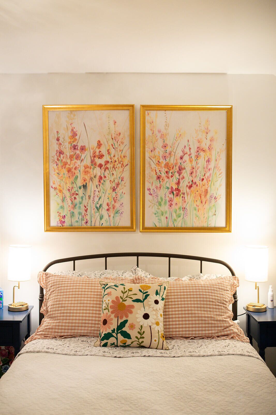 A bedroom with a bed, two framed floral paintings, a checkered pillow, and bedside lamps.