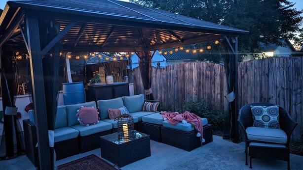 Un patio al aire libre con un área de estar cubierta, luces colgantes y una valla de madera al fondo.
