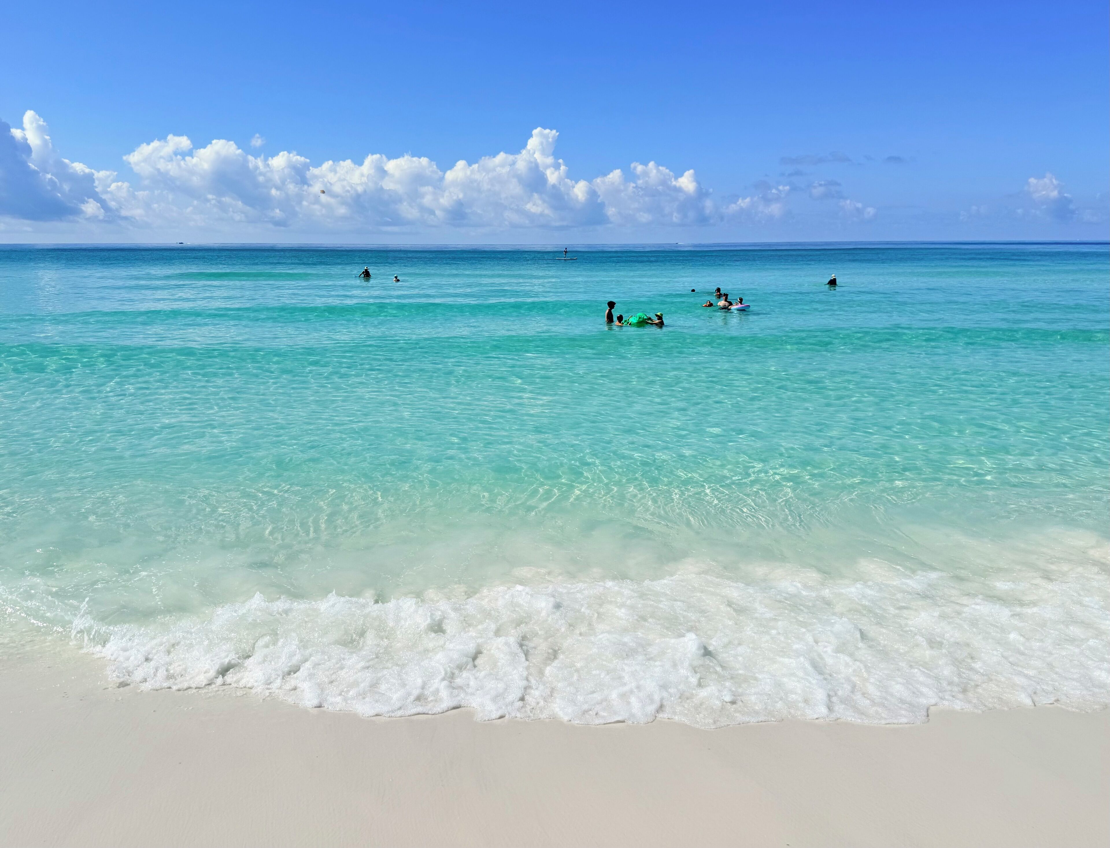 A beach with clear turquoise water, gentle waves, and a few people in the distance.