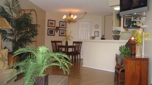 A living room with a wooden floor, a dining area with a table and chairs, a kitchen with a counter and cabinets, and a television mounted on the wall. -  Hidden Gem!  Exceptional 1 bed/1bath ocean & beachfront Kihei condo (Kihei)