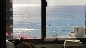Een kamer met een groot raam dat uitzicht biedt op zee, een bed met kussens, en een klein tafeltje met een mandje.
