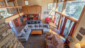 A spacious living area with a large sofa, a coffee table, and a dining table. There is a kitchen area with wooden cabinets and a stone wall. Large windows provide a view of the surrounding landscape. - Carson View, amazing views of Carson Peak and Gull Lake, walk to Gull Lake & town (June Lake)