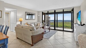 Een woonkamer met een bank, salontafel en televisie. Er is een glazen schuifdeur die leidt naar een balkon met uitzicht op de oceaan.