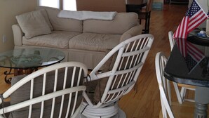 Una sala de estar con un sofá color beige, dos sillones de mimbre blanco, una mesa de centro de vidrio y una mesa de comedor con una bandera estadounidense. Hay ventiladores de techo, un mapa en la pared y un letrero que dice 'Welcome'.