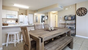 A dining area with a wooden table, chairs, and a bench. There is a kitchen area with white cabinets and appliances. A bookshelf with books is visible on the right.