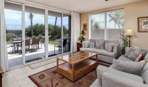 A living room with a sofa, a coffee table, and a sliding glass door leading to a patio with a table and chairs. - Island Princess #116 - 3Bd Poolside - Walkout to the Beach! (Fort Walton Beach)