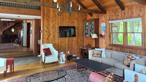 Amplia sala de estar con paredes de madera, una araña, un televisor, un mueble con puertas de vidrio y un ventanal con vistas a la vegetación.
