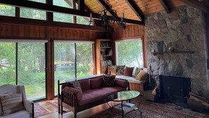 Un interior de cabaña acogedor, con techo de madera, ventanas amplias, chimenea de piedra y un rincón de estar cómodo.