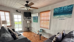 A living room with two grey sofas, a ceiling fan, a TV, and a painting of an underwater scene. - Seashell Cottage with breathtaking views! Near Swimming Pigs.  (exuma)