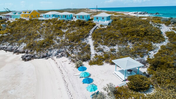Une propriété située sur la côte, avec plusieurs maisons aux couleurs vives, une plage de sable et un petit pavillon doté de parasols.