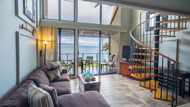A spacious living room with a curved staircase, a large sofa, and a view of the ocean through glass doors.
