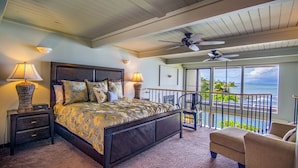 A bedroom with a bed, two nightstands, a chair, a balcony with a view of the ocean, and a ceiling fan.