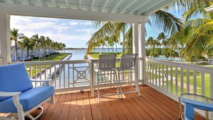 A covered outdoor area with a wooden deck, white railings, and a view of a water body and palm trees. - Waterfront luxury Villa (29) with sunset views and boat slip (Marathon)