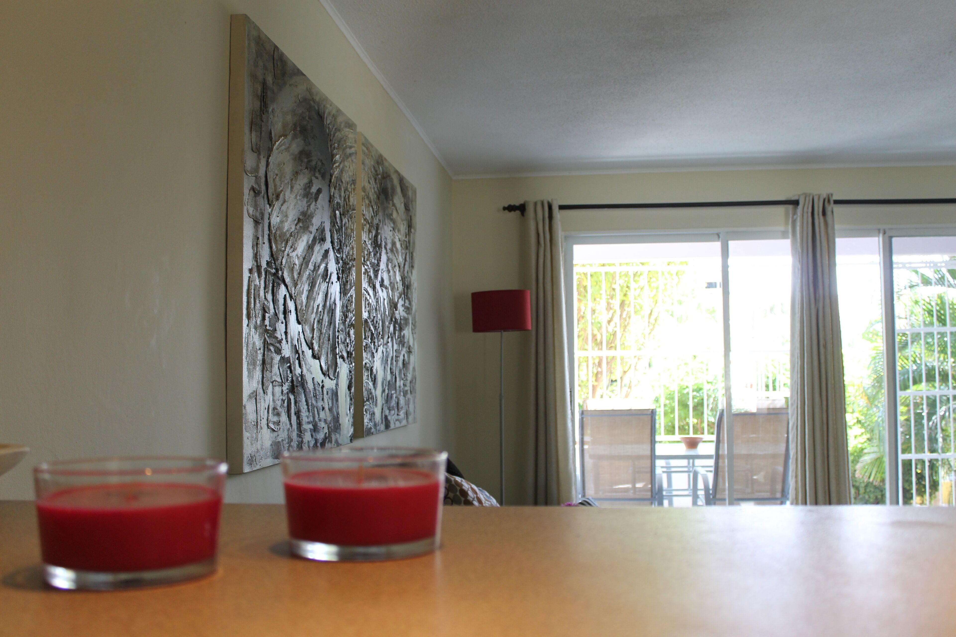 A room with a wooden table, two red candles, a large abstract painting, and a view of a balcony with outdoor furniture.