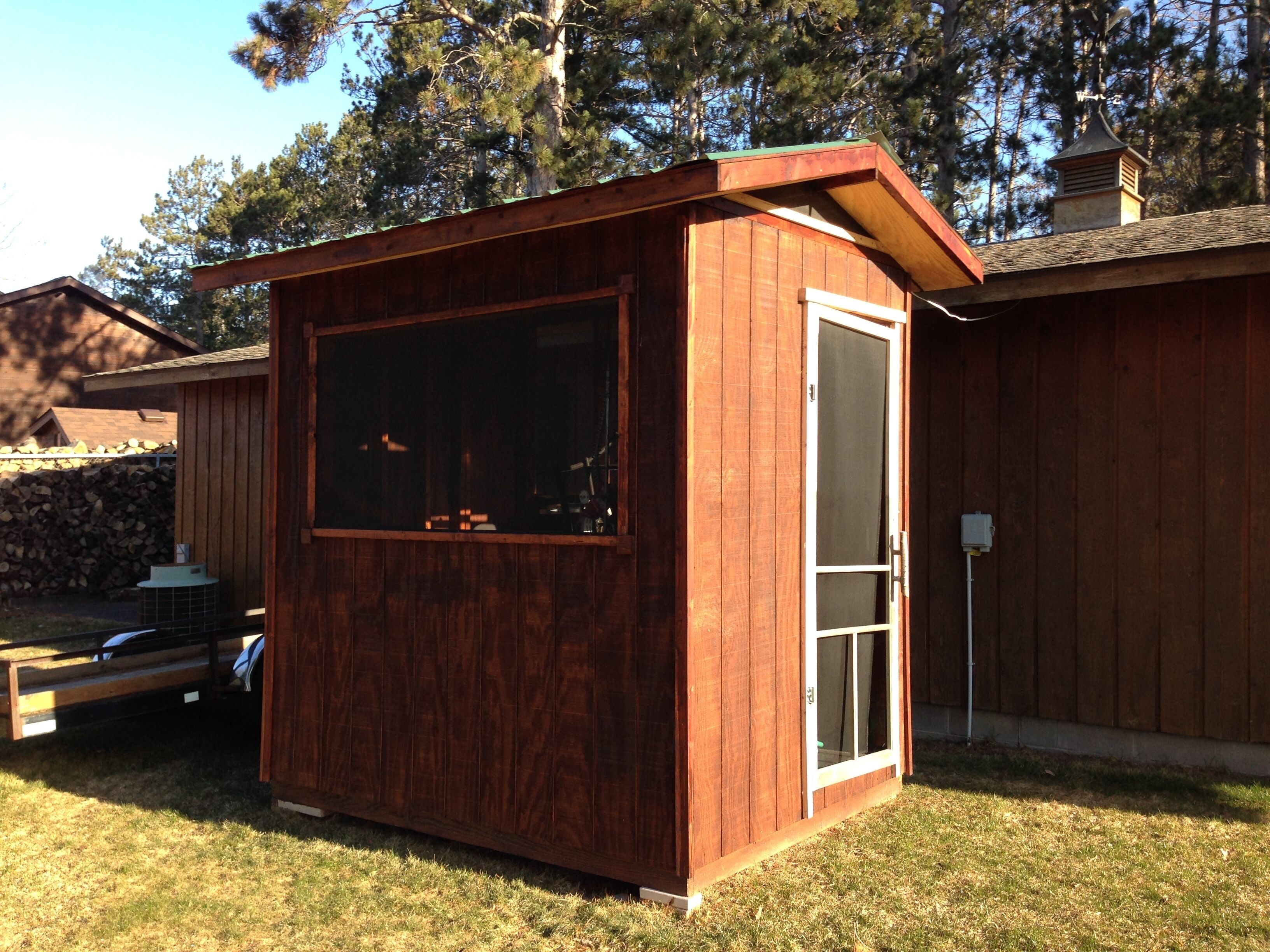 A small wooden cabin with a large window and a door, situated in a grassy yard with trees in the background.