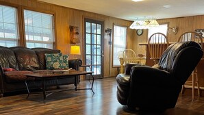 A living room with a wooden floor, a leather sofa, a black leather chair, and a dining table with chairs. - Beautiful Rustic Cabin On Lincoln Lake Is A Sportsman paradise.   (Kelso)