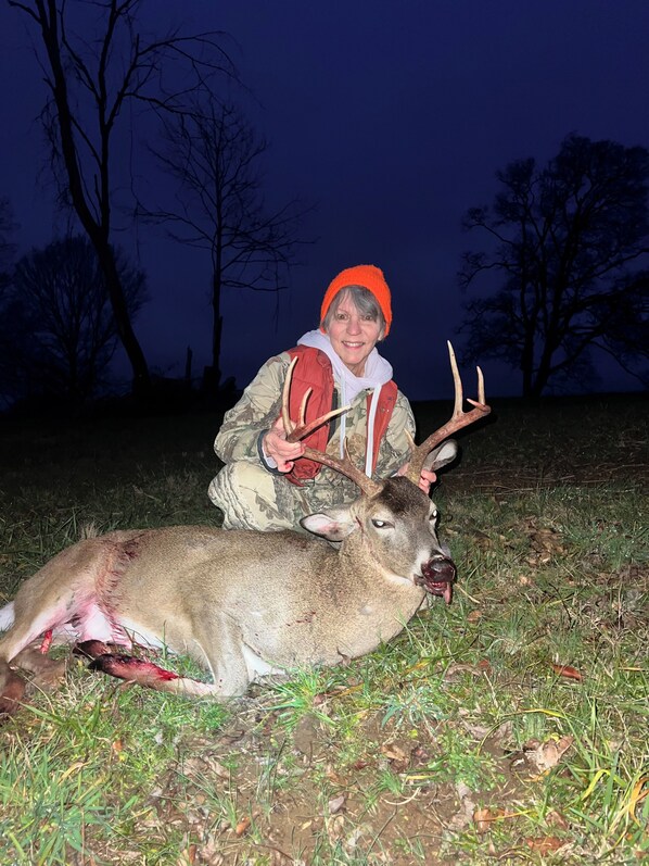 A person in camouflage attire holding antlers, standing next to a deer on grass.