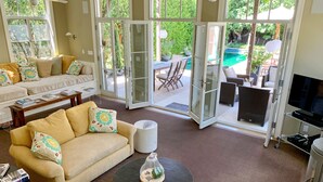 A spacious living room with a beige sofa, a coffee table, and a television. Large glass doors lead to an outdoor area with a pool and patio furniture. - Spa-Like Paradise Walking Distance To Wineries,  Shops, No Final Cleaning Fee (St. Helena)