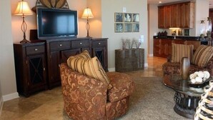 A living room with a sofa, ottoman, and a coffee table. There is a flat-screen TV on a stand, two lamps, and a decorative wall piece. - 360° View of Water - The Entire 18th Floor - 4 Bedroom Penthouse (Gulf Shores)