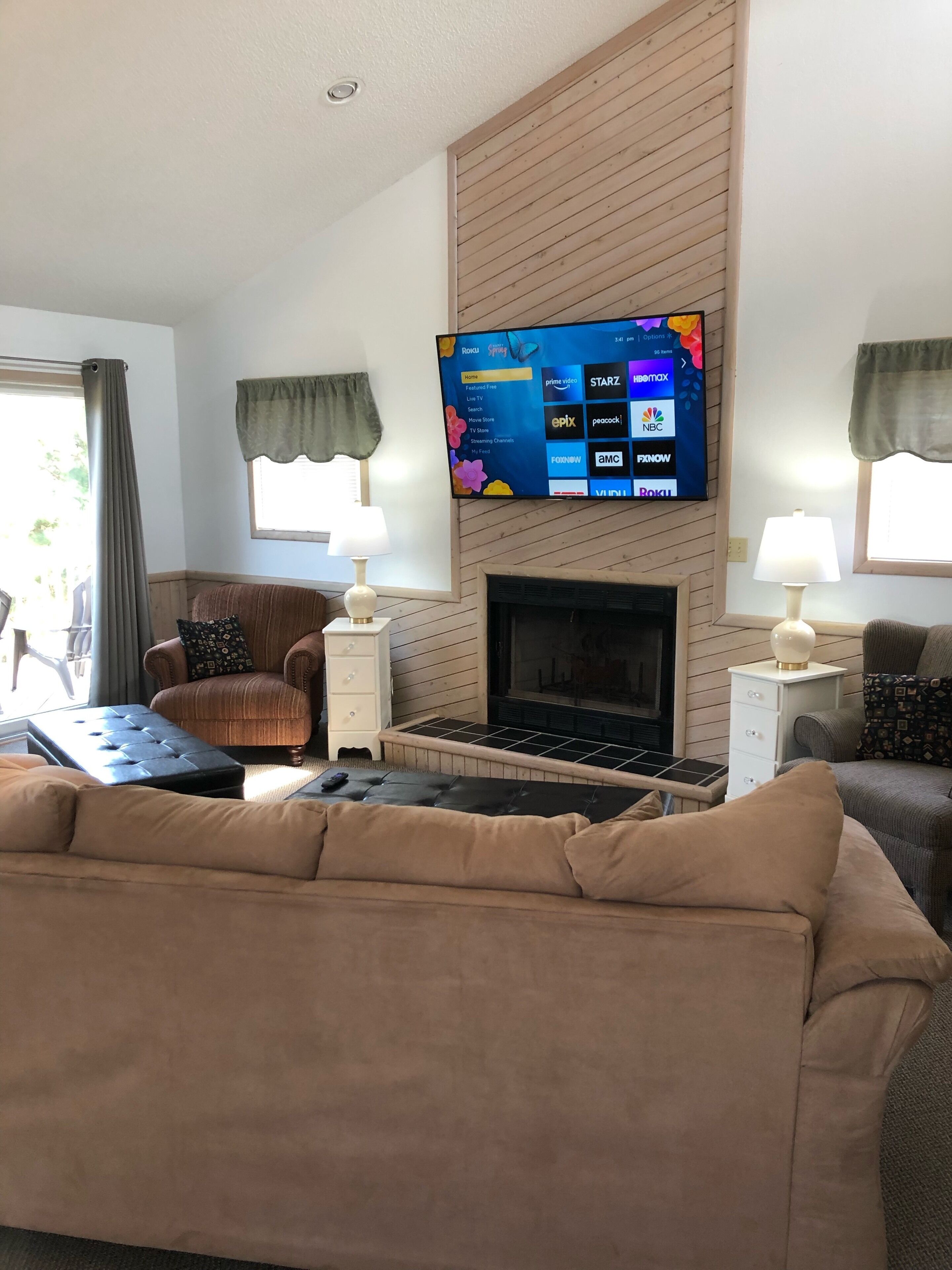 A living room with a flat-screen TV mounted above a fireplace, a beige sofa, a brown armchair, and two side tables with lamps.