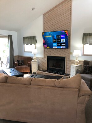 Sala de estar com televisão de tela plana acima de uma lareira, sofá bege, poltrona marrom e duas mesas laterais com luminárias.