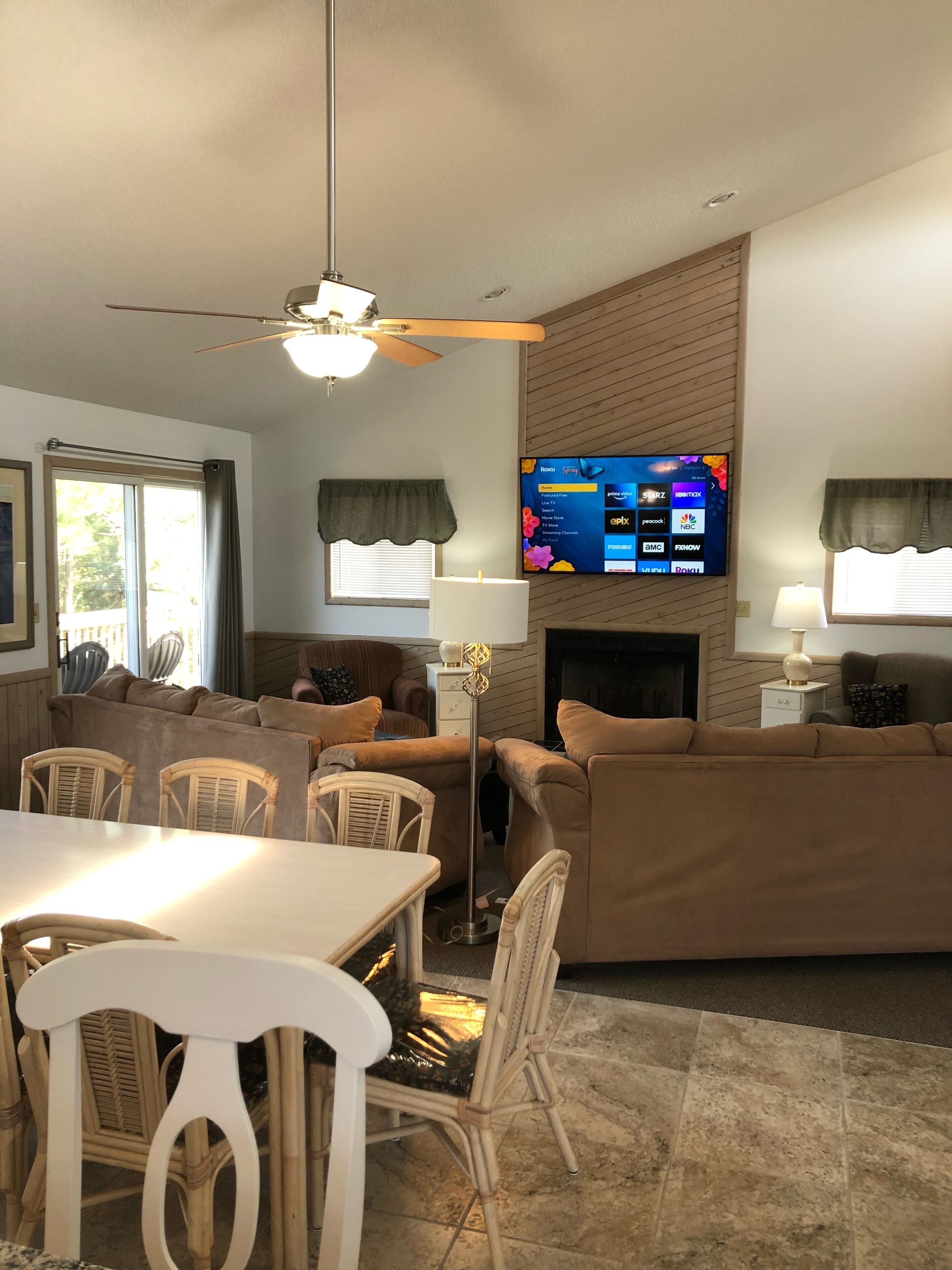 Ein Wohnzimmer mit an der Wand montiertem Fernseher, Deckenventilator, Kamin und Essbereich mit Stühlen und Tisch.