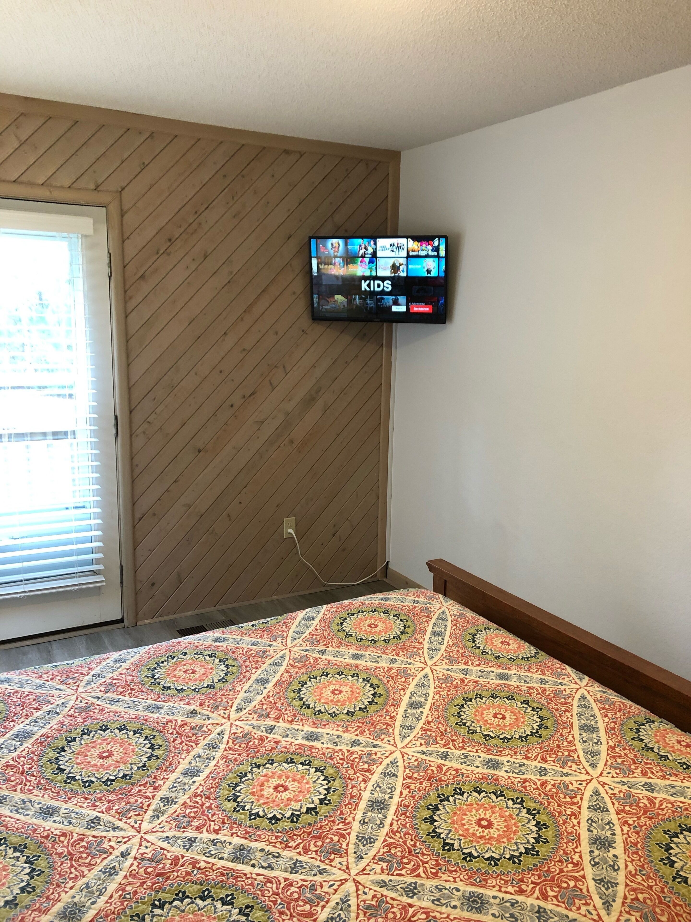 Een slaapkamer met een aan de muur bevestigde televisie, een bed met een decoratief sprei en houten lambrisering.