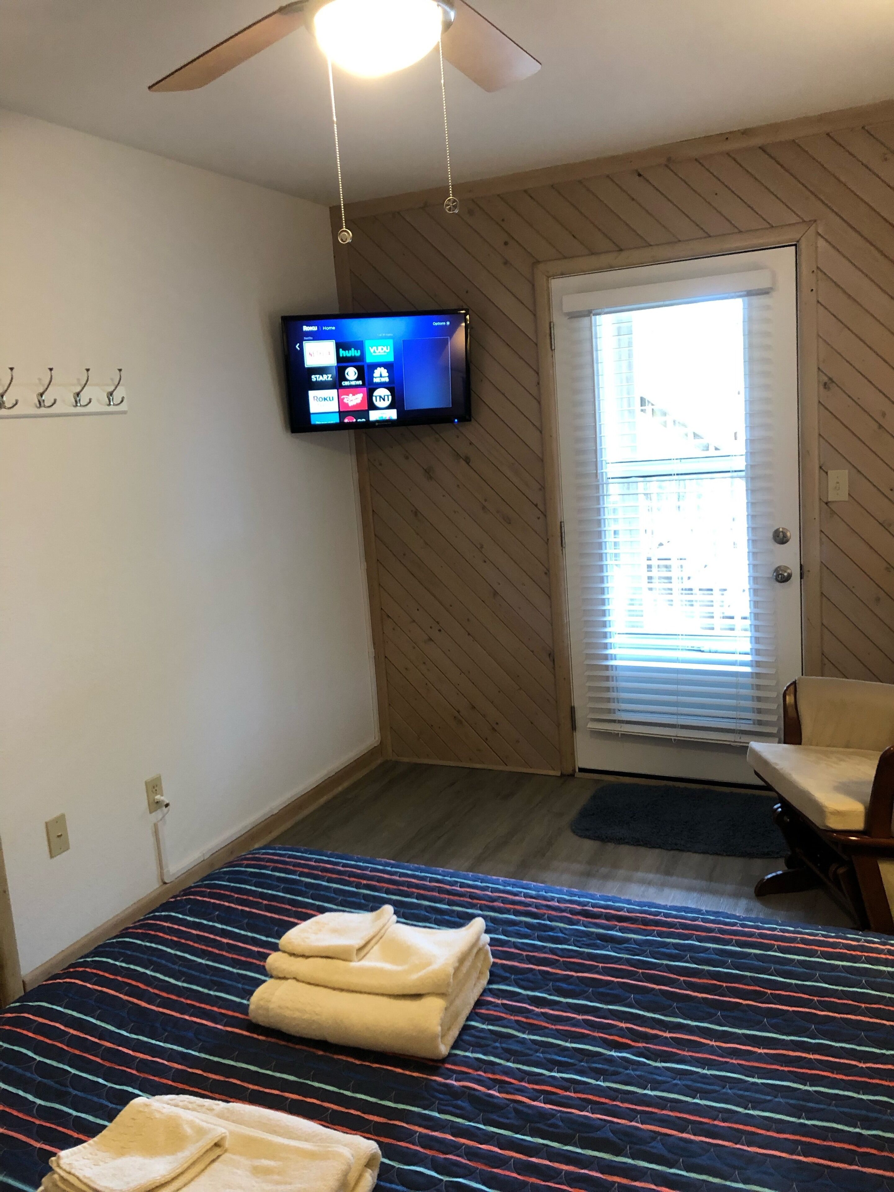 A bedroom with a bed, a television mounted on the wall, a door with blinds, and a ceiling fan.