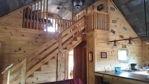 Interno di una baita in legno con una scala che conduce a una zona mansardata, una cucina con servizi essenziali e un arredamento rustico.