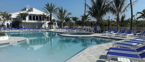 Zona de piscina con sillas reclinables azules, palmeras y un edificio al fondo.