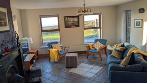 Ein gemütliches Wohnzimmer mit Kamin, einem blauen Sofa, einem Holztisch und zwei Sesseln. Große Fenster bieten Blick auf die Landschaft.