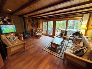A cozy living room with a fireplace, leather sofas, a television, and a large window with a view of trees. - Spa Cabin: Lakefront w Private Sauna & Hot Tub, Kayaks, Dock, 5 Star Clean Rated (Oakland)