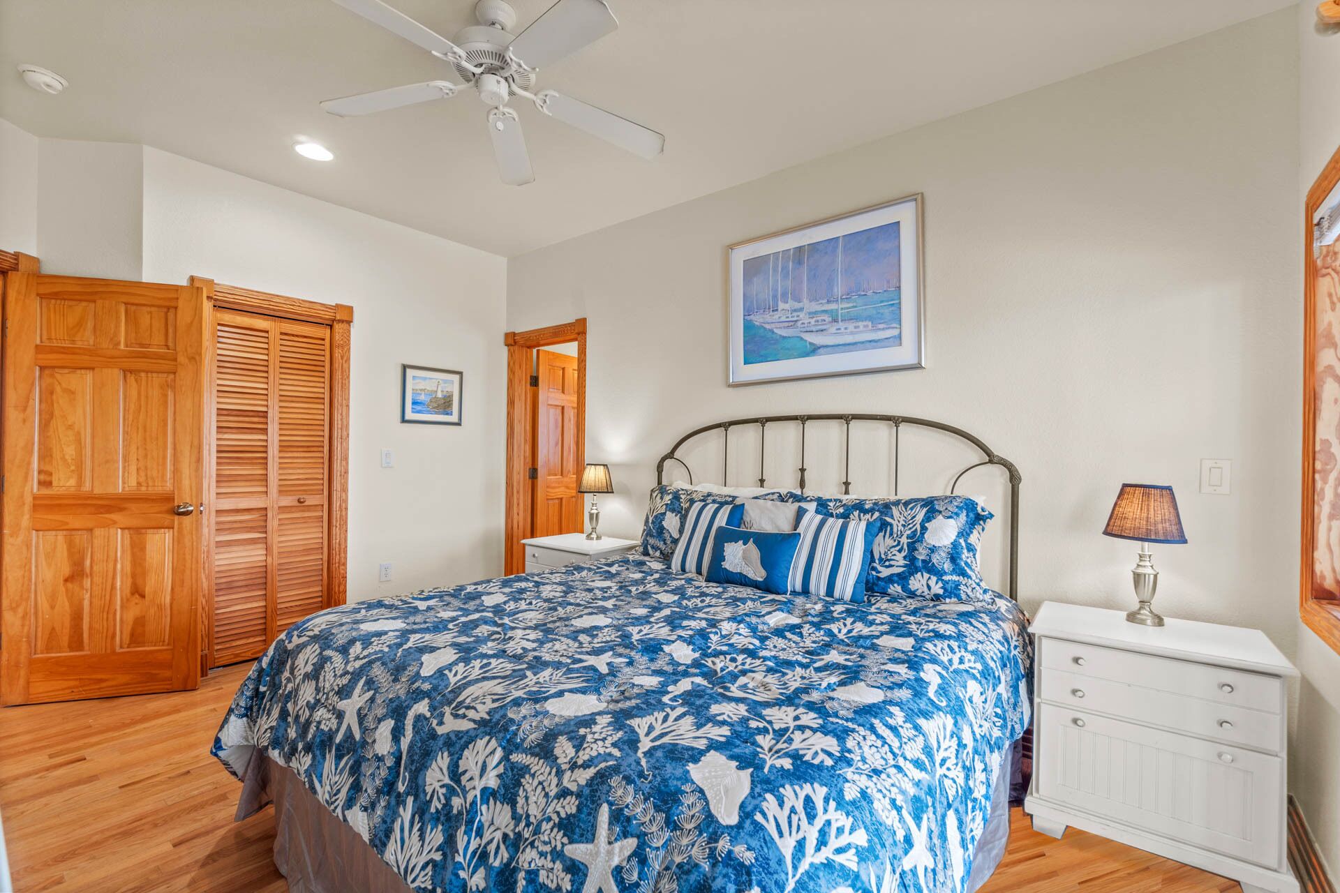 A bedroom with a bed, wooden doors, a ceiling fan, a nightstand, a lamp, and a framed picture on the wall.