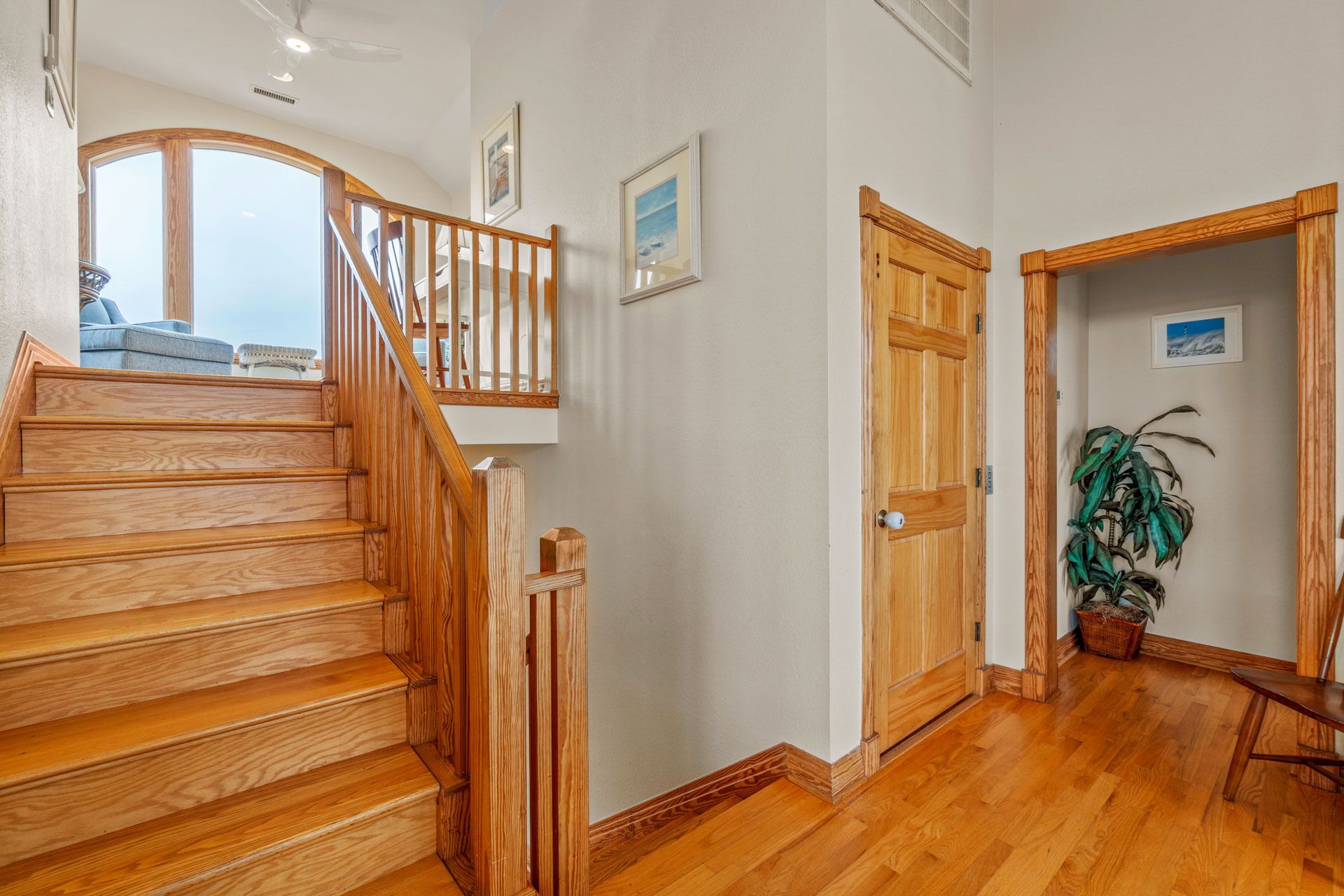 A wooden staircase leading to a balcony with a view, a door with a plant beside it, and a framed picture on the wall.