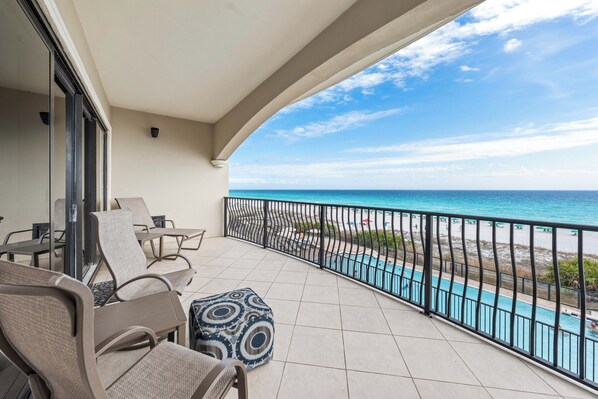 Ein Balkon mit Stühlen und einem gemusterten Hocker, mit Blick auf einen Pool und Strand.