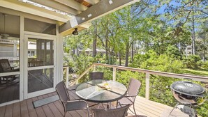 Eine überdachte Terrasse im Freien mit einem runden Glastisch, Korbstühlen, einem Grill und Blick auf eine Wohngegend mit Bäumen und Häusern.