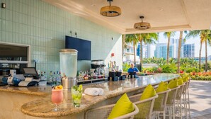 A bar area with a long counter, bar stools, a large TV screen, and a view of palm trees and buildings.