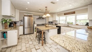Una cocina amplia con armarios blancos, un refrigerador de acero inoxidable, una isla central con taburetes y un letrero que dice 'WELCOME'.