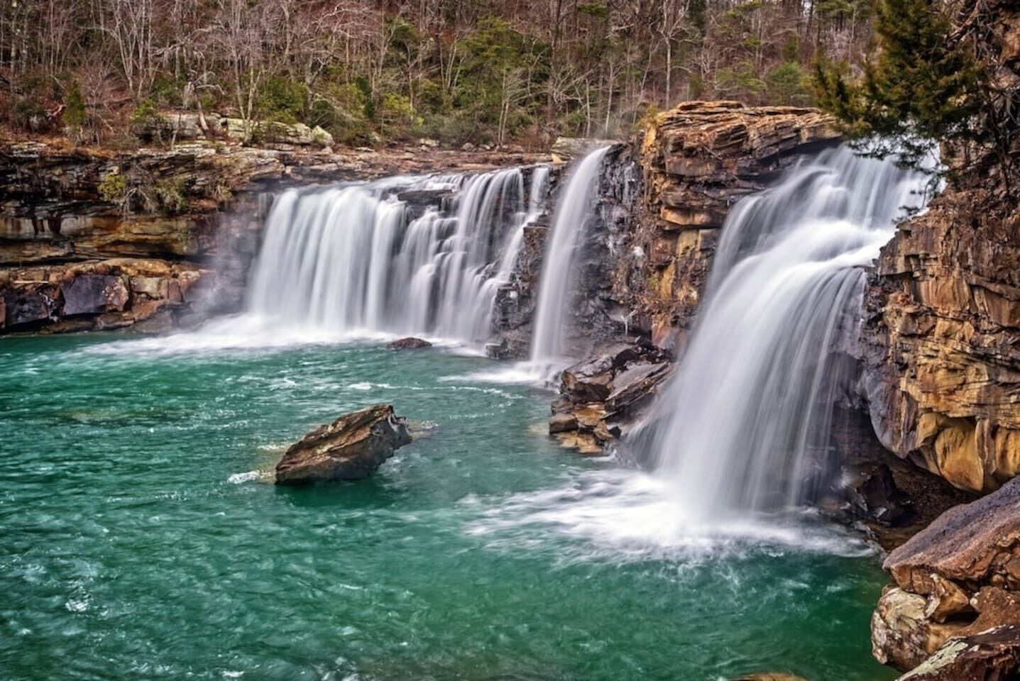 A waterfall cascading over rocky terrain into a clear turquoise pool surrounded by forested hills.
