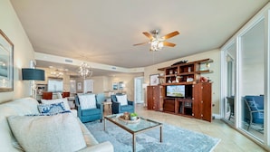 A spacious living area with a ceiling fan, a television, and a wooden entertainment center. - Caribe Resort D606 | Exquisite Bay Front Condo, Endless Amenities! (Orange Beach)