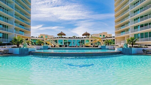 A hotel pool with a water slide, palm trees, and a clear blue sky. - Caribe Resort D606 | Exquisite Bay Front Condo, Endless Amenities! (Orange Beach)