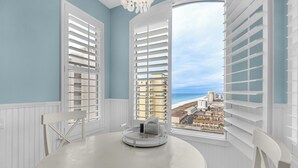 A room with a round table and a ceiling fan, large windows with white shutters, and a view of a beach and buildings. - Beach Colony West 16F- 3BR Gulf Front w/Beach Service and Onsite Management (Navarre)