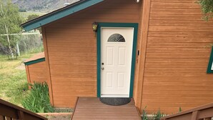 Un edificio de madera con una puerta blanca y detalles verdes, una terraza de madera que conduce a la entrada y un paisaje montañoso al fondo.